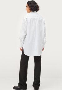 Witte katoenen shirt met lange mouwen, een knoopsluiting, achter plooidetail en licht gebogen zoom, gecombineerd met zwarte wijde broek.