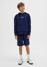 Completo con felpa blu navy e pantaloni corti realizzati in un mix di cotone, dotato di una tasca frontale e logo. Indossato con calzini bianchi e sneakers.