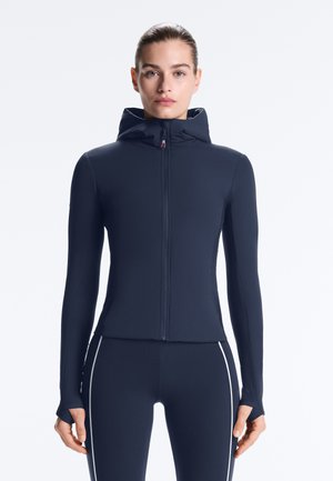 Femeie purtând un hanorac albastru închis cu fermoar și colanți sportivi asortați, cu dungi albe pe lateral, stând în picioare în fața unui fundal simplu.