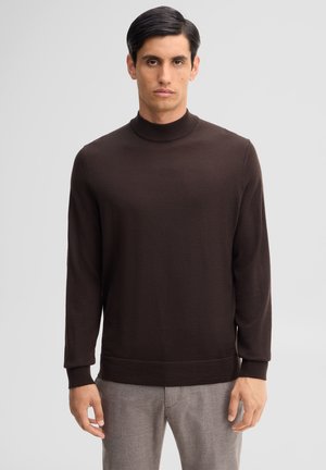 Uomo con capelli scuri che indossa un maglione a collo alto marrone scuro e pantaloni grigio chiaro, in piedi davanti a uno sfondo chiaro uniforme.