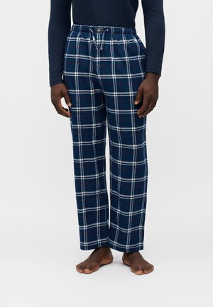 Mand iført marineblå og hvide ternede pyjamasbukser og en mørk langærmet trøje, stående barfodet mod en ensfarvet baggrund.