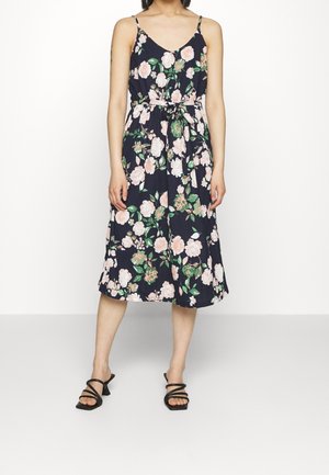 Kobieta w granatowej sukience midi w kwiaty w kolorze różowym i zielonym, z cienkimi ramiączkami, wiązaniem w talii oraz czarnych sandałach na obcasie z paskami.