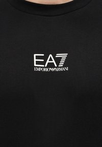 Tela negra con el logotipo blanco "EA7 Emporio Armani" centrado en la zona del pecho de una camiseta o sudadera.