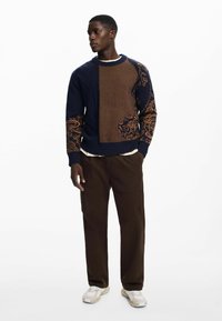 Navy sweater with textured orange diagonal stripe and decorative floral pattern on the sleeves; paired with brown pants and white sneakers.