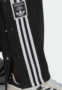 Black pants with white triple stripes and snap buttons, worn over white Adidas sneakers with black stripes and logo.