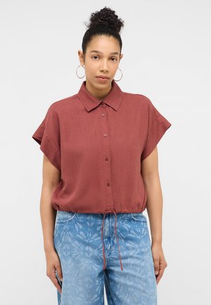 Woman with curly hair in a bun wearing hoop earrings, a rust-colored short-sleeve button-up shirt, and light blue patterned jeans.