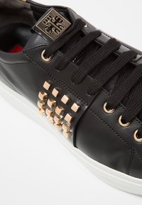 Baskets en cuir noir avec lacets noirs, clous métalliques dorés sur le côté et un emblème décoratif doré près du sommet des lacets.