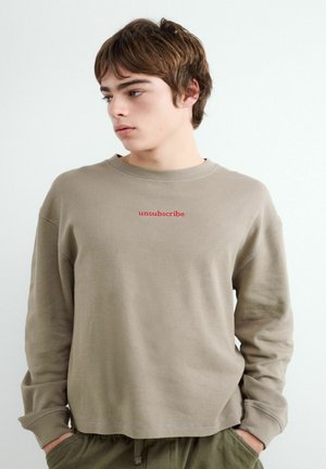 Mladá osoba s hnedými vlasmi v béžovej košeli s dlhým rukávom a červeným nápisom „odhlásiť sa“, ruky v vreckách, pozerá sa do strany.