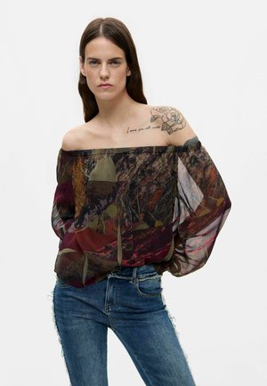 Vrouw met lang bruin haar draagt een off-shoulder, doorschijnende, patroon blouse en blauwe spijkerbroek, waarbij een bloemen tatoeage en tekst op de linker schouder zichtbaar zijn.