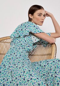 Fliederblaues Kleid mit floralem Muster in Grün, Blau und Weiß, mit Rüschensleeves und elastischem Bund, präsentiert vor einem geflochtenen Stuhl.