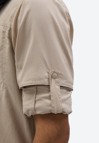 Bež košulja s kratkim rukavima, s rukavima podvrnutim i pričvršćenim tkaninom s gumbom na ruci osobe, na jednobojnoj pozadini.