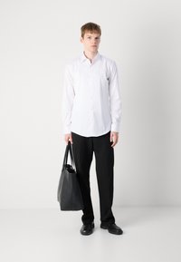 White button-up shirt with long sleeves, paired with black trousers and black leather shoes. Holding a black tote bag. Simple, structured styling.