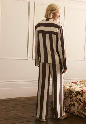 Kobieta stojąca w pomieszczeniu, ubrana w dopasowany beżowy i ciemnobrązowy pionowo paskowany top z długim rękawem i spodnie o szerokich nogawkach oraz kapcie.