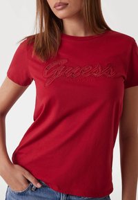 Raudonas medvilninis marškinėliai su išsiuvinėtu "Guess Los Angeles" logotipu išsiskiriančiu raštu, su klasikiniu apvaliu kaklu ir trumpomis rankovėmis.