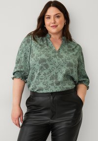Grøn bluse med blomsterprint, V-hals, korte pufærmer og struktureret stof, parret med sorte læderbukser med høj talje.