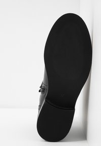 Chaussure en cuir noir avec une semelle texturée, de forme plate et un détail de boucle sur le côté. Étiquette de taille "37" visible sur la semelle.