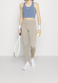 Sportovní outfit skládající se z modrého zkráceného topu a béžových legín, doplněný bílými teniskami, zápěstními pásky, teniskou a žlutým tenisovým míčkem.
