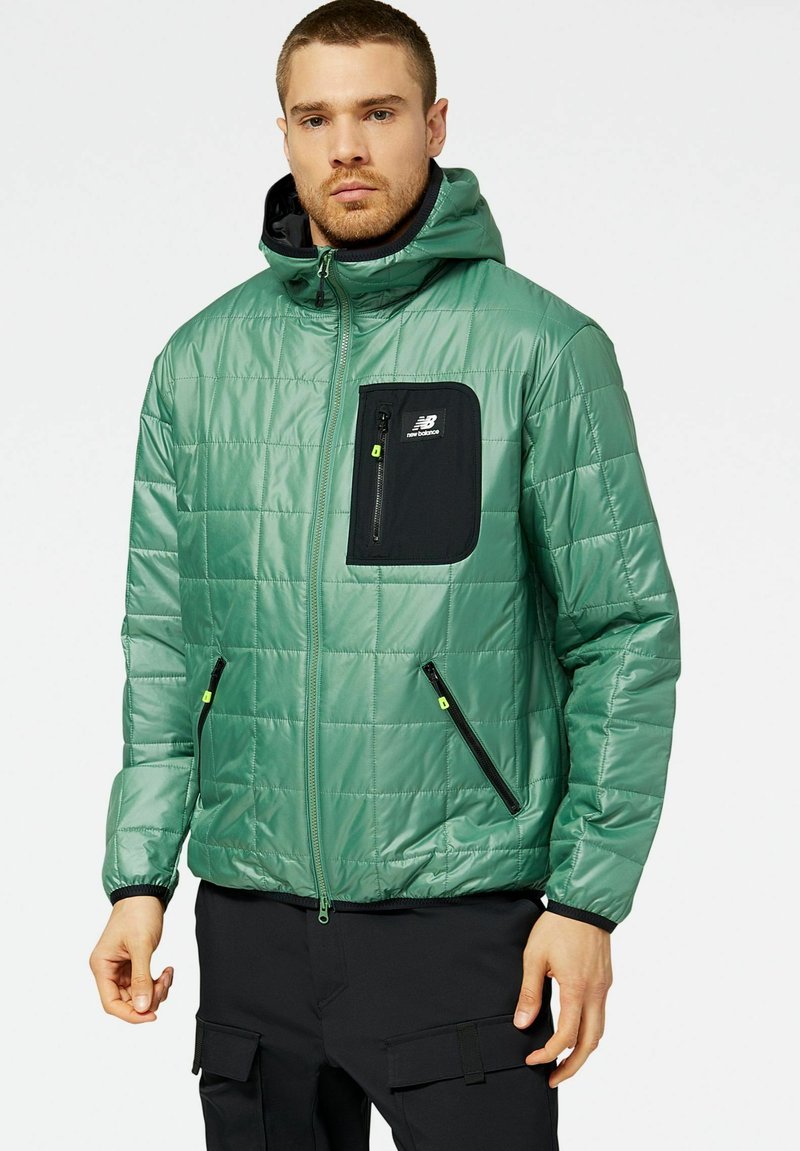 Mann in einer grünen Steppjacke mit Kapuze und schwarzen Reißverschlusstaschen, kombiniert mit schwarzen Cargo-Hosen.