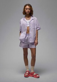 Camicia a maniche corte di colore viola chiaro con taschino e pantaloni corti abbinati; entrambi hanno una texture liscia. Indossato con sandali rosa e calzini beige.