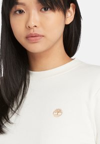 Vit bomullströja med en liten, broderad tanträdslogotyp på vänster bröstområde och ribbad rund hals.