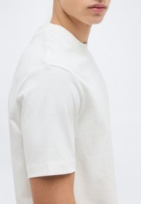 Νεαρό άτομο φορώντας απλό λευκό κοντομάνικο μπλουζάκι, σε κοντινή λήψη από τον ώμο μέχρι τα χείλη, πάνω σε ανοιχτό φόντο.