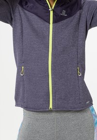 Lila idrottsjacka i texturerad väv, neon-gul dragkedja och två sidofickor. Svarta detaljer vid kragen förstärker designen.