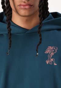 Tirkīzzila kapuce izgatavota no mīkstas auduma, ar rozā grafisko dizainu, kurā attēlota ceļgala figūra un logo uz priekšpuses, ar auklām un kapuci.