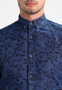 Sininen nappipaita, jossa on tummemman sinisiä lehtikuvioita, valmistettu denimin pehmeästä kankaasta ja siinä on perinteinen kaulus.