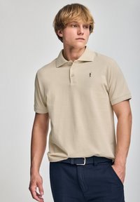 Camisa polo beige de manga corta, con tres botones y un pequeño logo negro. Combinada con pantalones azul marino oscuros y un cinturón trenzado.
