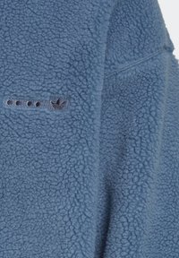 Blå fleecejacka med en strukturerad yta, som har en liten svart logotyp och knappdetaljer på framsidan. Mjuk, tjockt material.