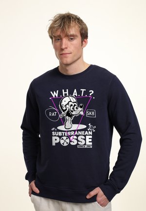 Jonge man met warrig blond haar die een marineblauwe trui draagt met een cartoonrat met een helm en de tekst "Subterranean Posse."
