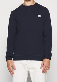 Uomo che indossa un felpa a maniche lunghe blu navy con un piccolo logo rettangolare bianco sul lato sinistro del petto e pantaloni color crema.