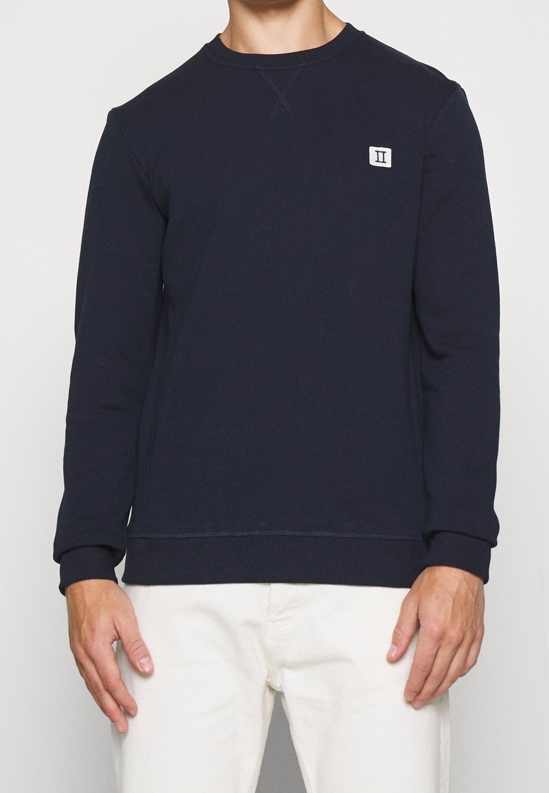 Uomo che indossa un felpa a maniche lunghe blu navy con un piccolo logo rettangolare bianco sul lato sinistro del petto e pantaloni color crema.