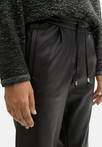 Schwarze, glatte Hose mit einem Bund mit Kordelzug und metallischen Akzenten. Kombiniert mit einem strukturierten, dunklen Strickpullover mit hellen Sprenkeln.