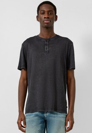 Mann trägt dunkelgraues Henley-Shirt mit kurzen Ärmeln und hellblaue Jeans, steht vor einfachem hellem Hintergrund.