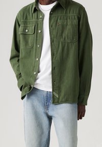 Camicia verde a maniche lunghe con chiusura a bottone, due tasche sul petto e un tessuto testurizzato. Indossata sopra una camicia bianca, abbinata a jeans azzurri chiari.