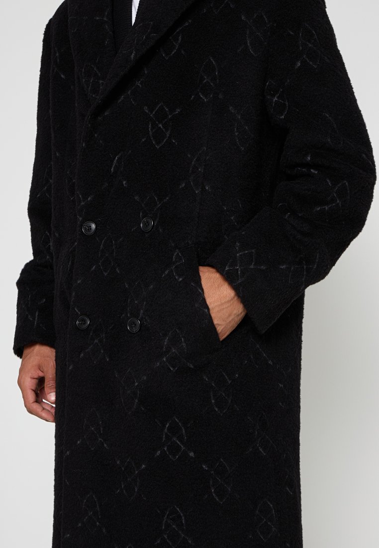 Manteau noir à double boutonnage avec un tissu texturé, présentant un motif subtil, deux boutons et des poches latérales. Manches longues et larges revers.