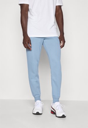 Pantaloni della tuta blu chiaro con elastico in vita e nelle caviglie, indossati con una maglietta bianca e sneakers bianche con accenti neri e rossi.
