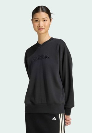 Joven mujer con sudadera negra de Adidas y falda negra con rayas blancas, de pie frente a un fondo gris claro.