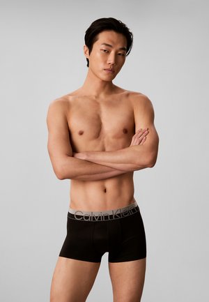 Hemdloser Mann mit verschränkten Armen, der schwarze Calvin Klein Boxershorts vor einem einfarbigen grauen Hintergrund trägt.