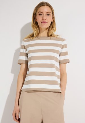 Jonge vrouw die een beige en wit horizontaal gestreept shirt met korte mouwen en beige broek draagt, staand tegen een effen witte achtergrond.