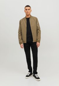 Olive green bomber jacket with a zippered pocket, paired with a black sweater and slim-fit black jeans. White laced sneakers complete the look.