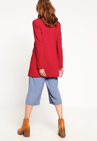 Röd lång blazer, lösa blå culotter och beige blockklackade ankelskorr. Blazern har strukturerade axlar och ett mjukt tyg.