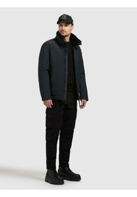 Czarna kurtka puchowa z wysokim kołnierzem, gładkim materiałem i zamkiem błyskawicznym. Połączona z czarnymi spodniami cargo i masywnymi czarnymi butami.