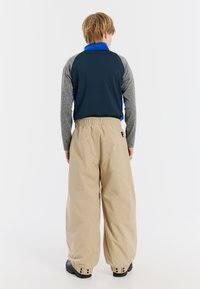 Jeugdoutfit met een donkerblauw en grijs langwerpig shirt met een opstaande kraag, gecombineerd met losse beige broeken en zwarte schoenen.