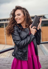 Black leather jacket, pink dress, holding a black wallet with white polka dots. Curly hair, smiling, outdoor urban setting.