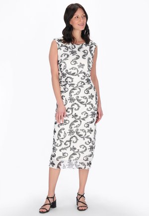 Kvinde iført en ærmeløs hvid midi-kjole med sorte blomsterkruseduller, sorte remsandaler med blokhæl, stående og kiggende til siden.