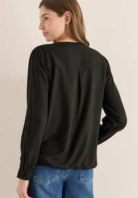 Schwarze Langarmbluse mit glatter Textur, geknöpften Manschetten und einer Falte am Rücken. Dazu getragen mit blauen Jeans.