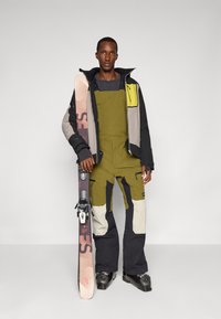 Casaco de ski com blocos de cor preto, cinza e verde-oliva, combinado com um fato de ski verde-oliva. O modelo segura um ski com um design rosa e preto.