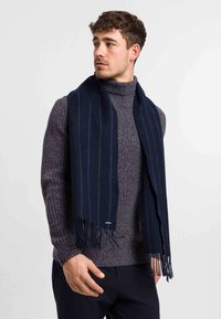 Sciarpa di lana blu marino con leggere righe verticali, dotata di bordi frangiati e una texture morbida, indossata sopra un maglione a collo alto grigio scuro a coste.